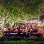 An outdoor event with dark long tables and hanging lights