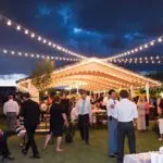 A group of people standing under a tent with bistro lighting.