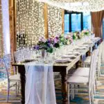 A long wooden table with white table cover and has clear flower vases with purple flowers