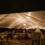 Event tent with tables and chairs under string lights, enhanced by professional lighting services.