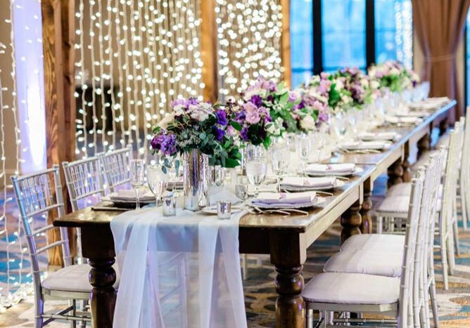 A long table set with flower vases with lavender flowers and clear vases with white lace table covers